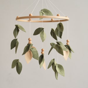Può includere: Un mobile in legno con foglie di feltro verde e perline di legno appese a una cornice circolare in legno. Il mobile è una decorazione per l'asilo nido.