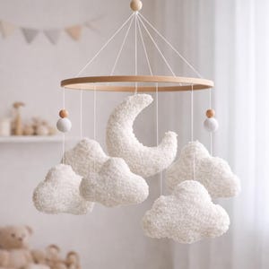 May include: A baby mobile featuring a wooden ring with hanging white clouds, a crescent moon, and wooden beads. The nursery decor is suspended above a white crib. The clouds and moon are made of a soft, textured material.