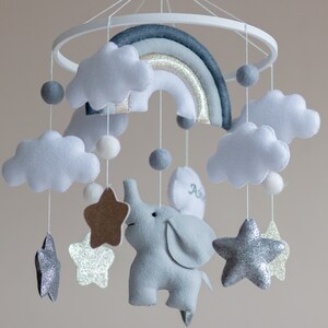 May include: A gray and white baby mobile with a rainbow, clouds, stars, and an elephant. The elephant is holding a star. The mobile is hanging from a white wooden ring.