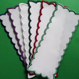 May include: A selection of white fabric bookmarks with scalloped edges, each trimmed with a different colour: purple, white, green, red, and green. The bookmarks are arranged in a fan shape against a green background.