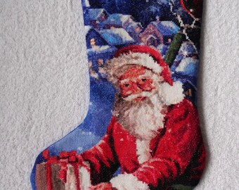 Könnte beinhalten: Weihnachtsstrumpf mit Santa Claus in rotem Anzug und Mütze, der ein Geschenk hält. Der Hintergrund zeigt ein verschneites Dorf in Blau- und Weißtönen. Der Strumpf hat einen grauen oberen Rand.