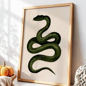 Puede incluir: Una lámina de arte mural vertical enmarcada que presenta una ilustración de una serpiente verde oscuro con el cuerpo enroscado y la lengua visible sobre un fondo de color crema sólido. La obra está en un marco de madera clara.