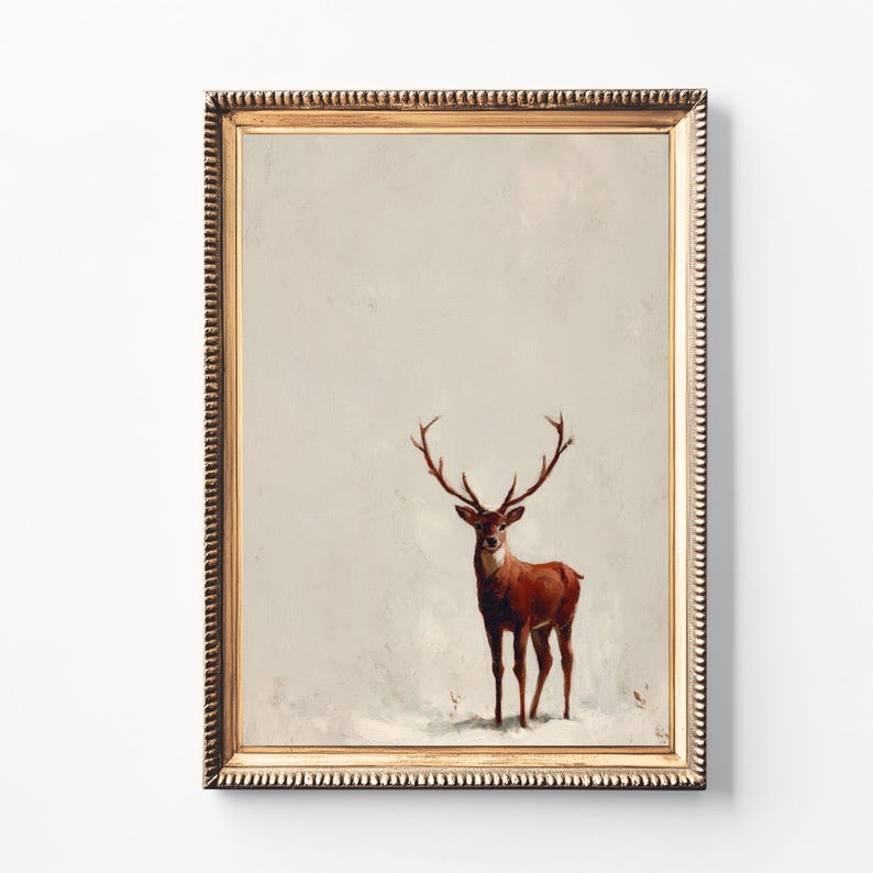 May include: A framed art print featuring a brown deer with large antlers standing in a snowy landscape. The artwork is set within a gold-colored, ornate frame, creating a vintage aesthetic. The background is a muted, off-white color.
