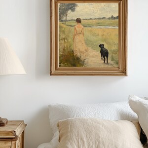 May include: A framed oil painting depicting a woman in a white dress walking with a black dog in a field. The painting is set against a white wall and a bed with white linens and a tan pillow.