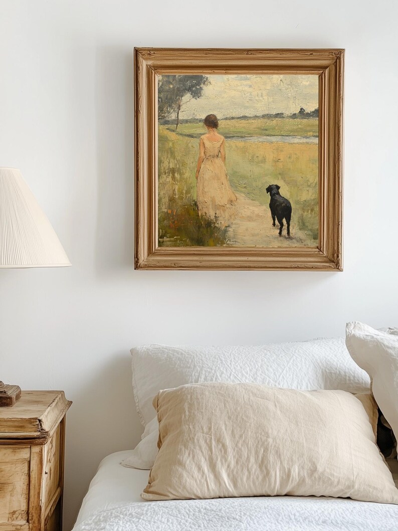 May include: A framed oil painting depicting a woman in a white dress walking with a black dog in a field. The painting is set against a white wall and a bed with white linens and a tan pillow.