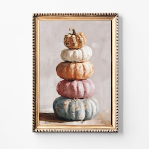 May include: A framed painting depicts a stack of pumpkins in shades of orange, white, pink, and blue. The pumpkins are arranged in a vertical tower, with the smallest pumpkin at the top. The frame is gold-colored.