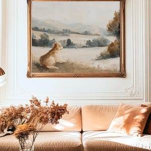 May include: An oil painting in a gold ornate frame depicts a golden retriever dog resting in a field, overlooking a landscape of rolling hills and trees. The painting hangs above a peach-colored sofa with matching pillows, and a glass vase with dried flowers.