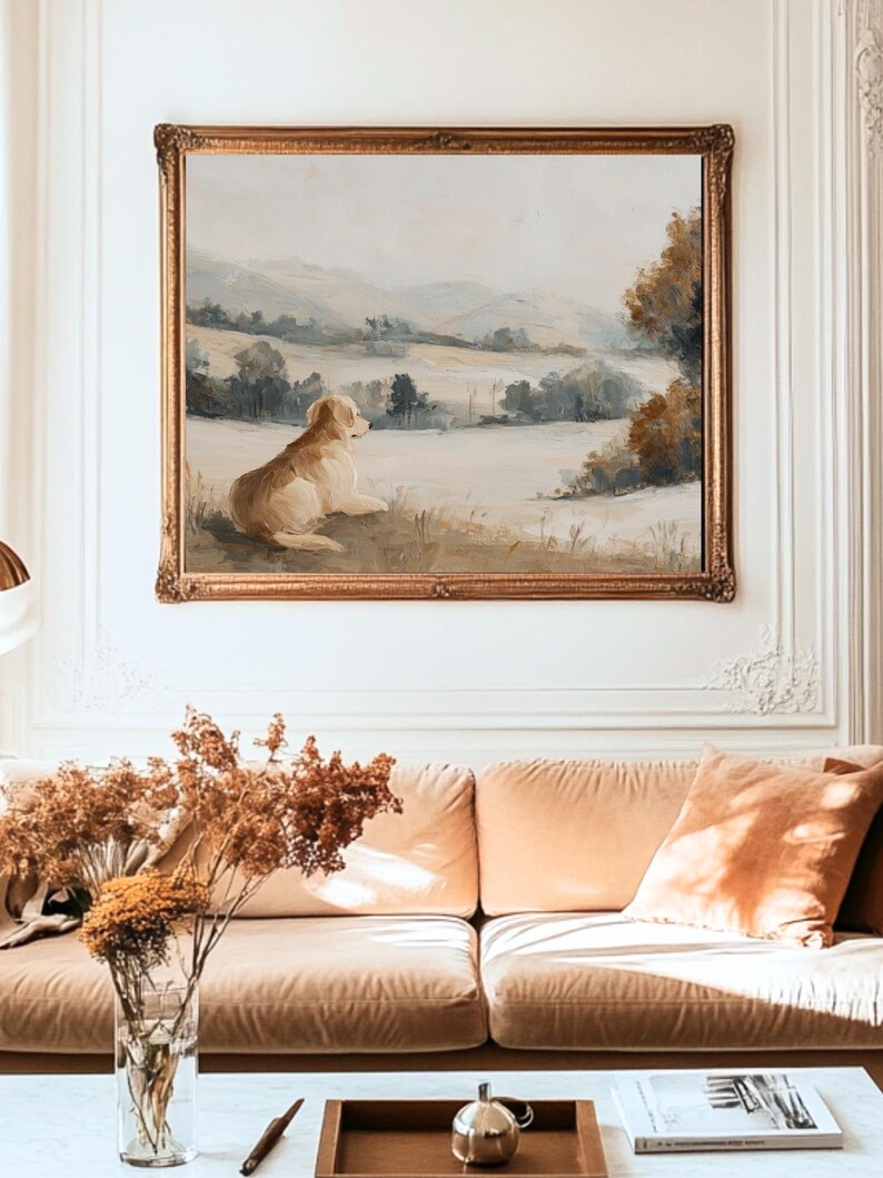 May include: An oil painting in a gold ornate frame depicts a golden retriever dog resting in a field, overlooking a landscape of rolling hills and trees. The painting hangs above a peach-colored sofa with matching pillows, and a glass vase with dried flowers.