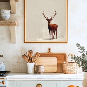 May include: A framed artwork featuring a brown deer with large antlers against a neutral background. The artwork is displayed in a kitchen setting, above a countertop with wooden cutting boards, utensils, and a basket. The overall aesthetic is rustic and warm.