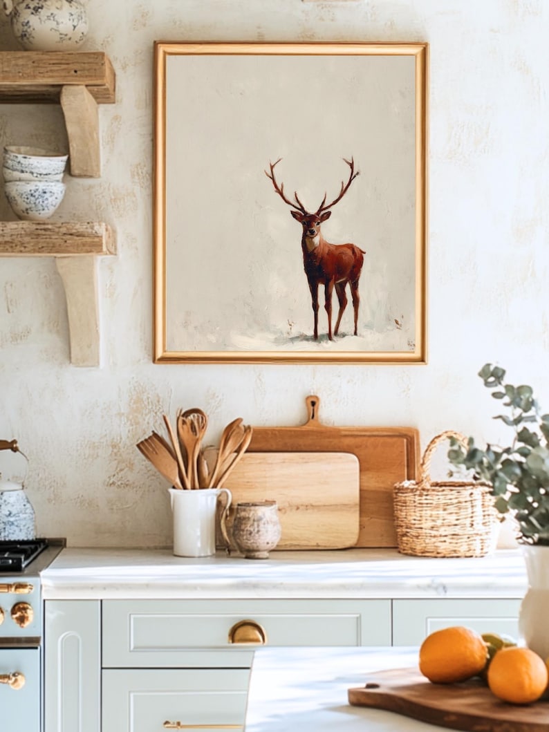 May include: A framed artwork featuring a brown deer with large antlers against a neutral background. The artwork is displayed in a kitchen setting, above a countertop with wooden cutting boards, utensils, and a basket. The overall aesthetic is rustic and warm.
