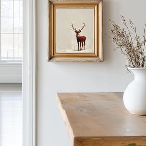 May include: A framed artwork featuring a deer in a snowy landscape. The deer is depicted in shades of brown and red, set against a neutral background. The artwork is encased in a gold-toned wooden frame, adding a touch of elegance to the piece.
