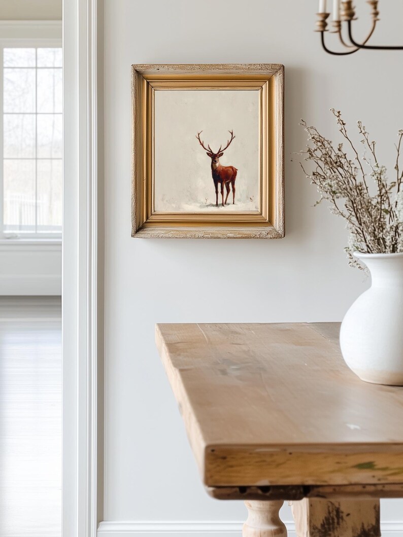 May include: A framed artwork featuring a deer in a snowy landscape. The deer is depicted in shades of brown and red, set against a neutral background. The artwork is encased in a gold-toned wooden frame, adding a touch of elegance to the piece.