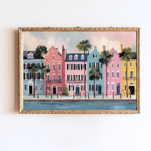 Puede incluir: Una impresión artística enmarcada que representa una fila de casas coloridas con palmeras. Las casas están pintadas en tonos de azul, rosa y amarillo, con contraventanas y puertas negras. La obra de arte está ambientada contra un cielo pastel y una vía fluvial azul.