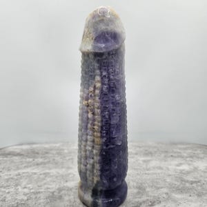 May include: A carved stone sculpture in the shape of a phallus. The sculpture is primarily purple with areas of white and tan. The surface has a textured pattern. The sculpture is standing upright on a gray surface.