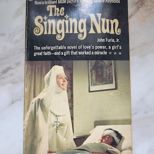 May include: Vintage paperback book cover for "The Singing Nun" by John Furia, Jr. The cover features a nun in a white habit and a patient in a hospital bed. The title is in large, stylized letters. The cover also includes text about the novel and the MGM picture starring Debbie Reynolds.