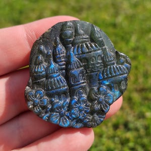 May include: A carved, oval-shaped labradorite stone featuring a detailed relief of a cityscape with domes and floral accents. The stone displays iridescent blue hues, with dark blue and black tones. The carving is held in a hand against a blurred green background.