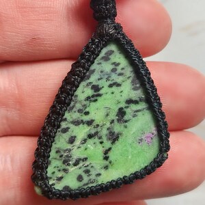Peut inclure: Un pendentif triangulaire compos&eacute; d'une pierre verte tachet&eacute;e de noir avec une petite inclusion magenta, ench&acirc;ss&eacute; dans un sertissage en cordon macram&eacute; noir avec une boucle nou&eacute;e au sommet, pr&eacute;sentant une bordure en fibre tress&eacute;e &agrave; la main.