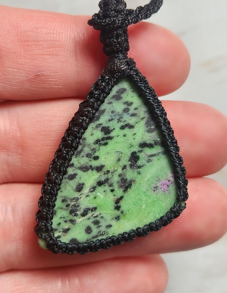 Peut inclure: Un pendentif triangulaire compos&eacute; d'une pierre verte tachet&eacute;e de noir avec une petite inclusion magenta, ench&acirc;ss&eacute; dans un sertissage en cordon macram&eacute; noir avec une boucle nou&eacute;e au sommet, pr&eacute;sentant une bordure en fibre tress&eacute;e &agrave; la main.