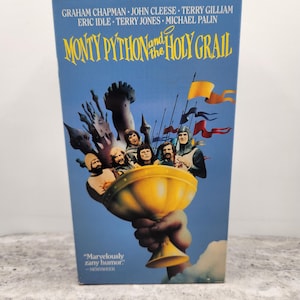 May include: A blue VHS tape case for the film Monty Python and the Holy Grail. The cover features a large golden chalice held by a hand, with several figures and a castle silhouette above it. Text includes the cast names and the quote Marvelously zany humor.