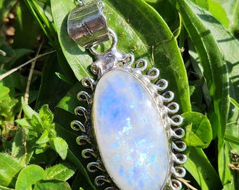 May include: A silver pendant necklace featuring an oval moonstone. The moonstone has a blue and white iridescent appearance, set in a decorative silver frame with a delicate chain. The necklace is displayed on a bed of green grass.