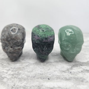 May include: Three small carved stone skulls, one gray, one green, and one green with pink and black markings.