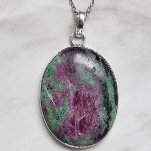 May include: An oval-shaped pendant featuring a polished ruby zoisite gemstone with vibrant magenta and deep green inclusions, set in a simple silver-toned metal bezel. The pendant hangs from a delicate silver-toned chain with a classic link design.