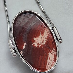 Peut inclure: Collier pendentif ovale avec une pierre polie brun-rouge&acirc;tre vein&eacute;e de blanc, sertie dans un cadre en m&eacute;tal argent&eacute;. Le pendentif est suspendu &agrave; une cha&icirc;ne serpent argent&eacute;e, cr&eacute;ant un accessoire simple mais &eacute;l&eacute;gant.