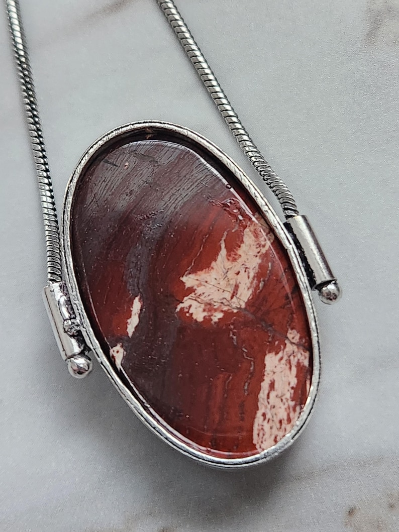 Peut inclure: Collier pendentif ovale avec une pierre polie brun-rouge&acirc;tre vein&eacute;e de blanc, sertie dans un cadre en m&eacute;tal argent&eacute;. Le pendentif est suspendu &agrave; une cha&icirc;ne serpent argent&eacute;e, cr&eacute;ant un accessoire simple mais &eacute;l&eacute;gant.