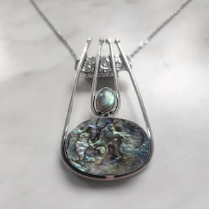 May include: A silver pendant necklace featuring a large, oval-shaped abalone shell inlay with iridescent blue, green, and purple tones. Above the shell sits a small, round, greyish-blue labradorite gemstone, all framed by four vertical silver prongs.