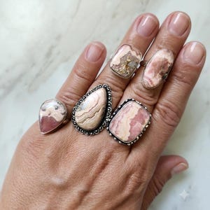 May include: Four silver rings featuring polished, banded pink and cream-colored stones. The rings include a teardrop shape, a large pear shape with a beaded border, a rectangular stone, and an oval stone, all set in sterling silver metal bands.