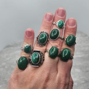 Pode incluir: Uma m&atilde;o usando oito an&eacute;is de prata com pedras ovaladas de malaquita verde. Os an&eacute;is s&atilde;o de v&aacute;rios estilos e tamanhos.
