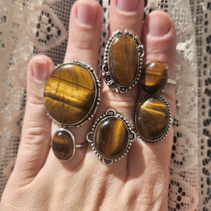 May include: Five silver rings featuring oval tiger's eye gemstones. The rings have elaborate silver detailing around the stones.