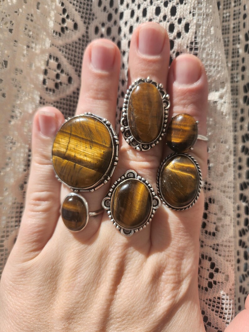 May include: Five silver rings featuring oval tiger's eye gemstones. The rings have elaborate silver detailing around the stones.
