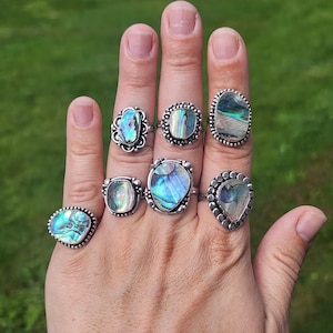 May include: A collection of seven silver rings featuring iridescent abalone shell inlays. Each ring displays unique, swirling patterns of blue, green, and purple hues set within textured or beaded silver metal bands of varying shapes and sizes.