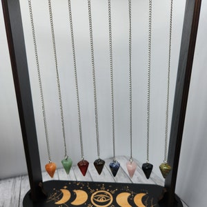 May include: A wooden stand with a black base and gold accents displays nine crystal pendulums on silver chains. The pendulums are made of various gemstones, including red, green, orange, brown, blue, pink, and clear.