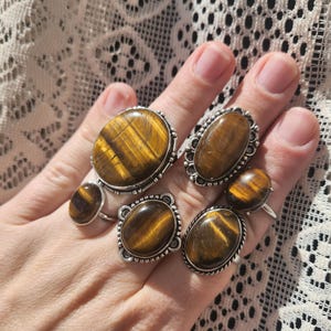 May include: Set of five silver rings featuring oval-shaped tiger's eye gemstones. The rings have a silver band with a decorative border around the gemstone.