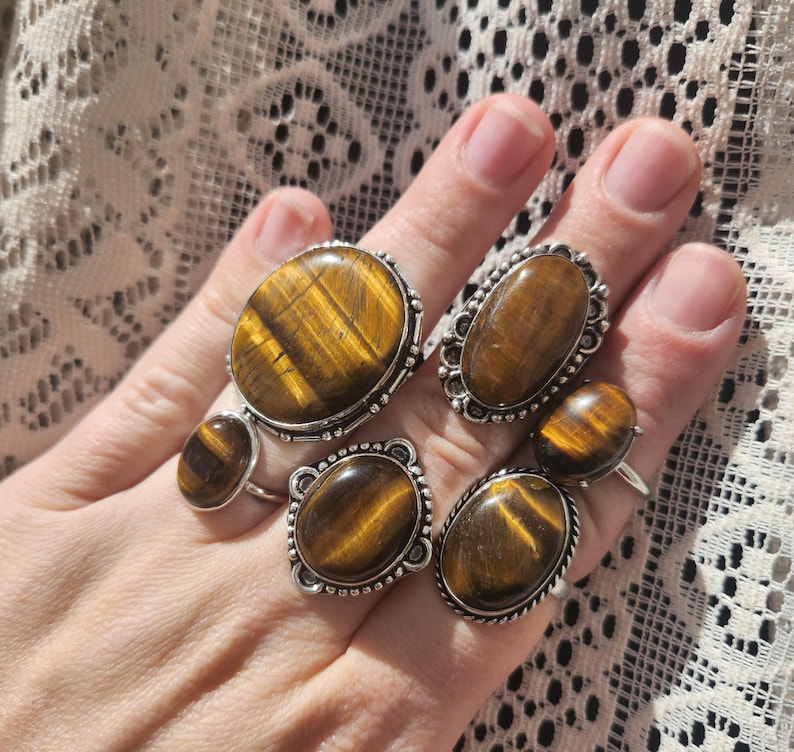 May include: Set of five silver rings featuring oval-shaped tiger's eye gemstones. The rings have a silver band with a decorative border around the gemstone.
