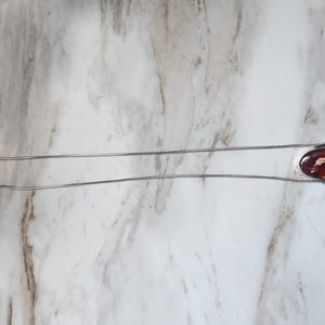 Peut inclure: Un collier argent&eacute; avec un pendentif ovale. Le pendentif pr&eacute;sente un motif rouge fonc&eacute; et blanc. La cha&icirc;ne est de type serpentin fin. Le fermoir est visible. Le collier mesure environ 45 cm de long.