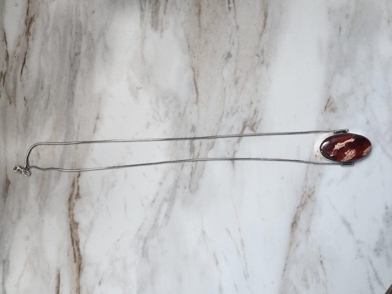 Peut inclure: Un collier argent&eacute; avec un pendentif ovale. Le pendentif pr&eacute;sente un motif rouge fonc&eacute; et blanc. La cha&icirc;ne est de type serpentin fin. Le fermoir est visible. Le collier mesure environ 45 cm de long.