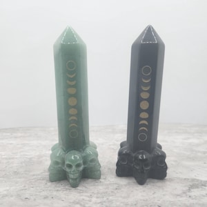 May include: Two crystal wands with a skull base and moon phases etched on the front. One wand is green and the other is black.