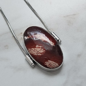 Peut inclure: Un collier argent&eacute; avec un pendentif ovale. Le pendentif pr&eacute;sente une pierre brun-rouge&acirc;tre polie avec des veines blanches et cr&egrave;me. La pierre est sertie dans une lunette argent&eacute;e et suspendue &agrave; une fine cha&icirc;ne argent&eacute;e.