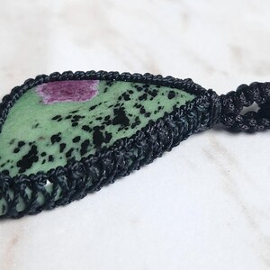 Peut inclure: Un pendentif compos&eacute; d'une pierre de zo&iuml;site triangulaire verte avec des taches noires et une inclusion magenta, ench&acirc;ss&eacute;e dans un sertissage en macram&eacute; noir avec une attache de collier en cordon tress&eacute; assorti, montrant des n&oelig;uds complexes.