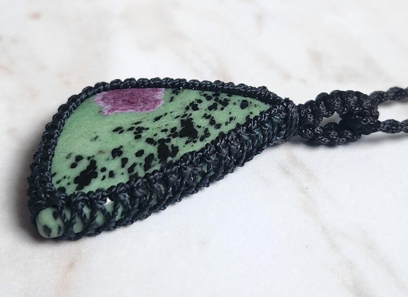 Peut inclure: Un pendentif compos&eacute; d'une pierre de zo&iuml;site triangulaire verte avec des taches noires et une inclusion magenta, ench&acirc;ss&eacute;e dans un sertissage en macram&eacute; noir avec une attache de collier en cordon tress&eacute; assorti, montrant des n&oelig;uds complexes.