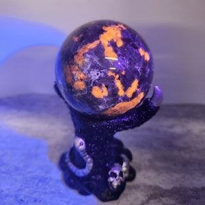 May include: A dark purple and orange crystal sphere sits on a black, skeletal hand stand. The sphere is illuminated with a bright, orange glow.