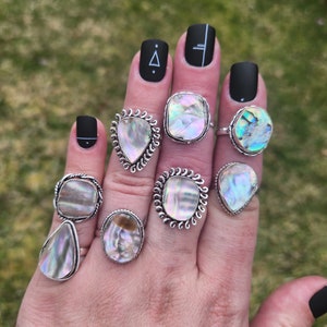 May include: A hand wearing seven silver rings with iridescent abalone shell inlays. The rings are on different fingers and are all different shapes and sizes.