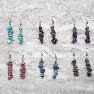 May include: A collection of dangle earrings with various natural stone beads in shades of yellow, turquoise, brown, purple, blue, green, and pink. Each earring features a silver-colored hook and a vertical arrangement of small, irregular-shaped stones.
