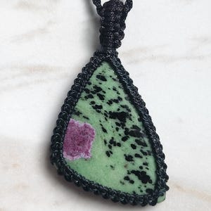 May include: A triangular pendant featuring a green stone with black speckles and a distinct magenta inclusion, encased in a black macrame cord setting. The pendant hangs from a matching black braided cord necklace with a knotted macrame bail.