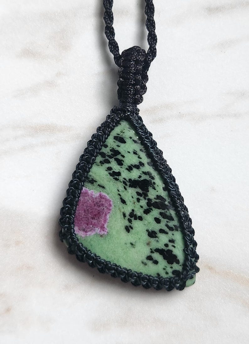 Peut inclure: Un pendentif triangulaire compos&eacute; d'une pierre verte tachet&eacute;e de noir avec une inclusion magenta, serti dans un cordon en macram&eacute; noir. Le pendentif est suspendu &agrave; un collier en cordon tress&eacute; noir assorti avec une b&eacute;li&egrave;re en macram&eacute;.