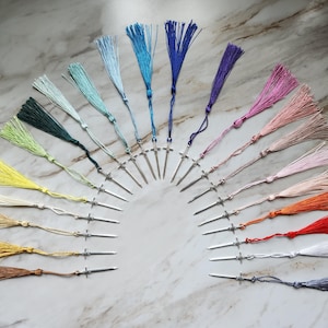 May include: A collection of colorful tassel bookmarks arranged in a semi-circle. Each bookmark features a silver-toned metal pin with a decorative tassel in shades of blue, green, yellow, pink, orange, and brown. The background is a marble-like surface.
