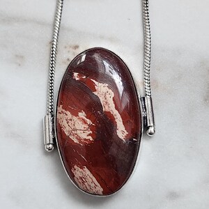 Peut inclure: Un collier argent&eacute; avec un pendentif ovale. Le pendentif pr&eacute;sente une pierre polie brun-rouge&acirc;tre avec des veines blanches et cr&egrave;me. La pierre est sertie dans une lunette argent&eacute;e et suspendue &agrave; une cha&icirc;ne argent&eacute;e.